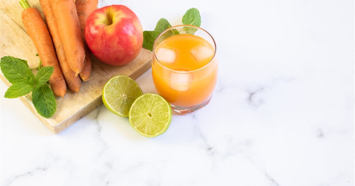 Glass of fruit smoothie with ice beside carrots, an apple, mint and limes on a wooden board