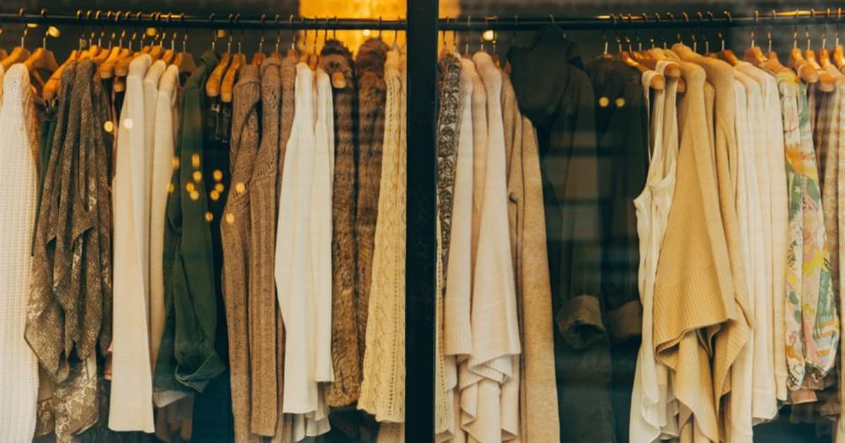 Rail of knitwear and jackets in cream, olive and tan hanging behind a lit boutique window
